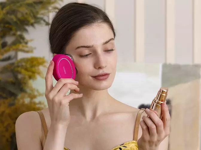 A young woman with sleek dark hair in yellow dress using FOREO BEAR 2 microcurrent device and conductive SUPERCHARGED serum 